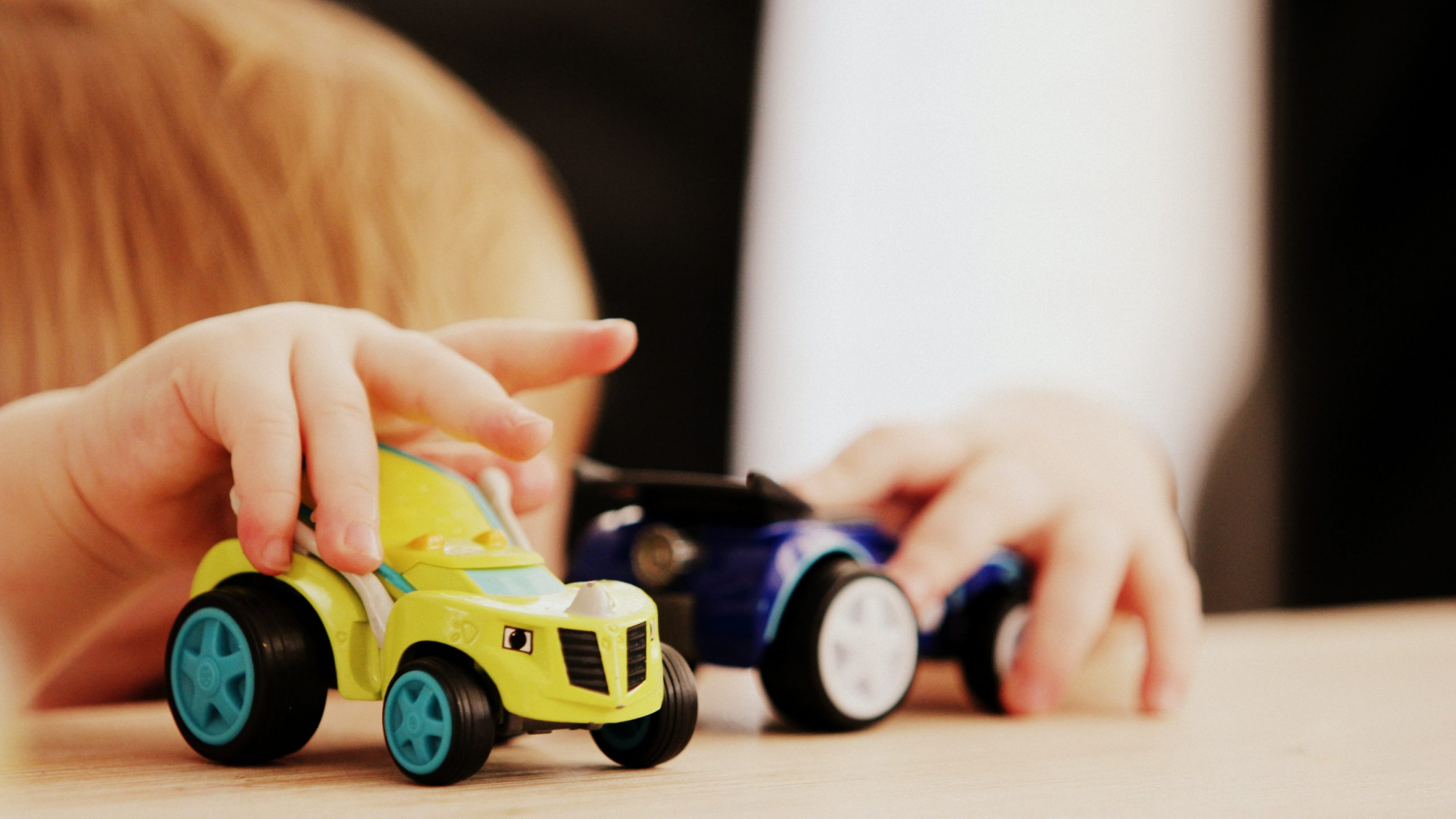 child playing with two assorted-color car plastic toys on brown wooden table