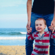 man assisting baby to walk on beach